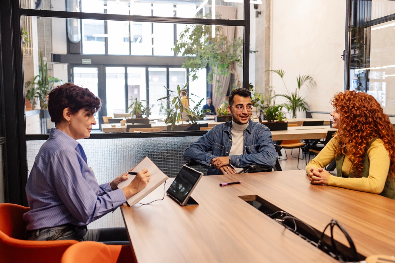 Human resources manager is taking notes while interviewing two young smiling candidates sitting at a desk in a modern office with plants and glass walls